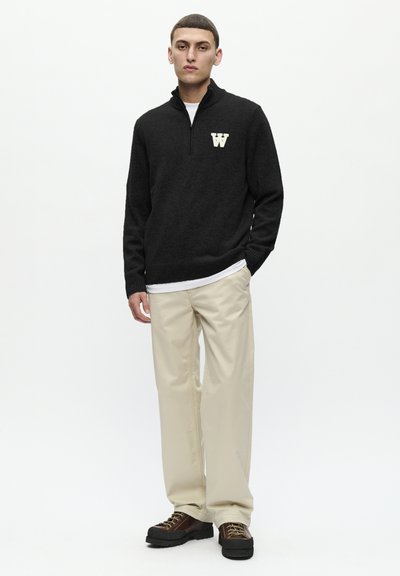 Un pull en tricot noir avec un col montant et un logo "W" blanc sur la poitrine, accompagné d'un pantalon beige et de chaussures à lacets marron foncé.