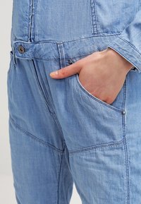 Ljust blå denimoverall med en framficka, dekorativ sömnad och metallknappdetaljer. Har en avslappnad passform och textilier med struktur.