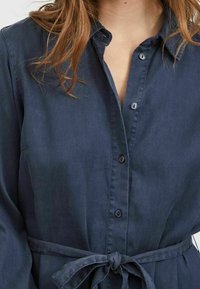 Mörkblå skjortklänning i slät tyg, med knappframsida, krage och midjeband. Långa ärmar med manschettdetaljer.