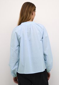 Lichtblauwe blouse met een gerimpelde hals, lange mouwen en broderie anglaise accenten op de manchetten. Zacht, lichtgewicht katoen.