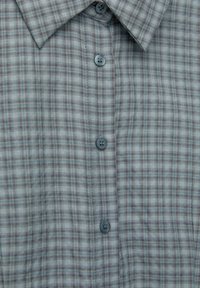 Chemise à boutons avec un motif à carreaux gris clair, agrémentée d'accents bleus et bordeaux. Le tissu présente une finition douce et texturée avec cinq boutons.