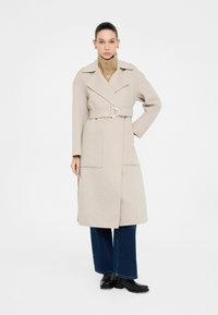 Cappotto in lana beige con vita a cintura, colletto a punta, tasche laterali e maniche lunghe, abbinato a jeans ampi blu scuro e stivali neri.