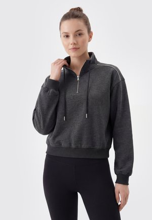 Ung kvinde iført en mørkegrå halv-zip sweatshirt og sorte leggings, holder i lynlåsen tæt ved halsen mod en ensfarvet baggrund.