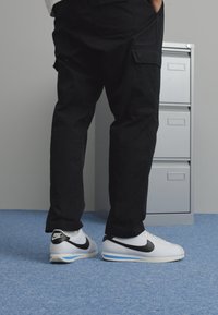 Fehér Nike sportcipők fekete swoosh-sal, texturált talppal, fekete cargo nadrággal párosítva. A háttérben szürke irattárolók és kék szőnyeg található.