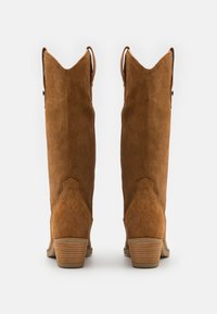 Botas de ante marrón hasta la rodilla con una punta afilada, acentos de cortes angulares y un tacón apilado. Presentan una textura suave y detalles de costura.