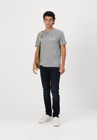 Grå bomullst-skjorte med hvit "TOMMY HILFIGER" logo, kombinert med mørkeblå skinny jeans og hvite sneakers. Modellen holder en lys brun veske.