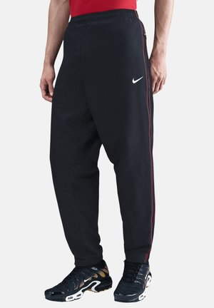 Mann trägt schwarze Nike Trainingshose mit roten Seitenstreifen und schwarze Nike Sneakers, steht vor hellem Hintergrund.