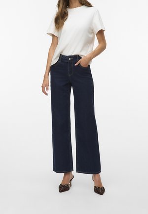 Mujer con camiseta blanca lisa de manga corta, vaqueros anchos oscuros y tacones marrones de punta con detalles decorativos.