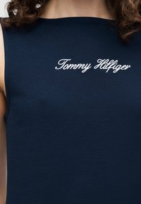 Σκούρο μπλε αμάνικο τοπ με λευκό κεντημένο λογότυπο «Tommy Hilfiger» στο πάνω μέρος του στήθους, φορεμένο από άτομο με σγουρά μαλλιά.
