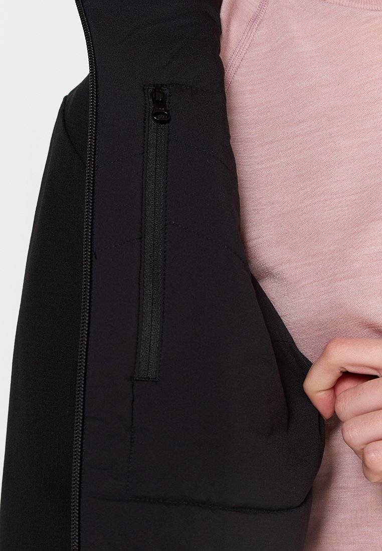 Chaqueta negra aislante con bolsillo con cremallera, con textura acolchada y tela suave, superpuesta a una camisa rosa clara.