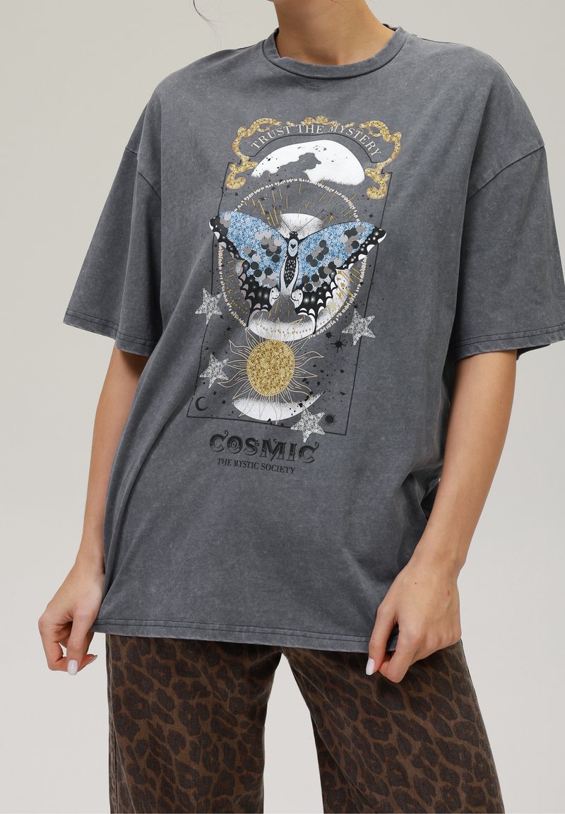 T-shirt gris avec un grand papillon bleu, lune, soleil, étoiles et texte "Trust the Mystery" et "Cosmic The Mystic Society." Pantalon léopard marron.