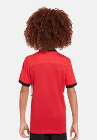 Maglia sportiva rossa con maniche corte, colletto nero e un tessuto texturizzato e traspirante con dettagli sui pannelli laterali.