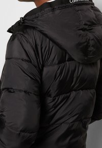 Giacca a piumino nera con cappuccio, design trapuntato e texture lucida. Presenta il marchio Calvin Klein sul colletto. Leggera e isolante.