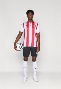 Jogador de futebol masculino está em pé segurando uma bola de futebol, vestindo uma camisola listrada em vermelho e branco, calções pretos, meias brancas e chuteiras brancas, em fundo simples.