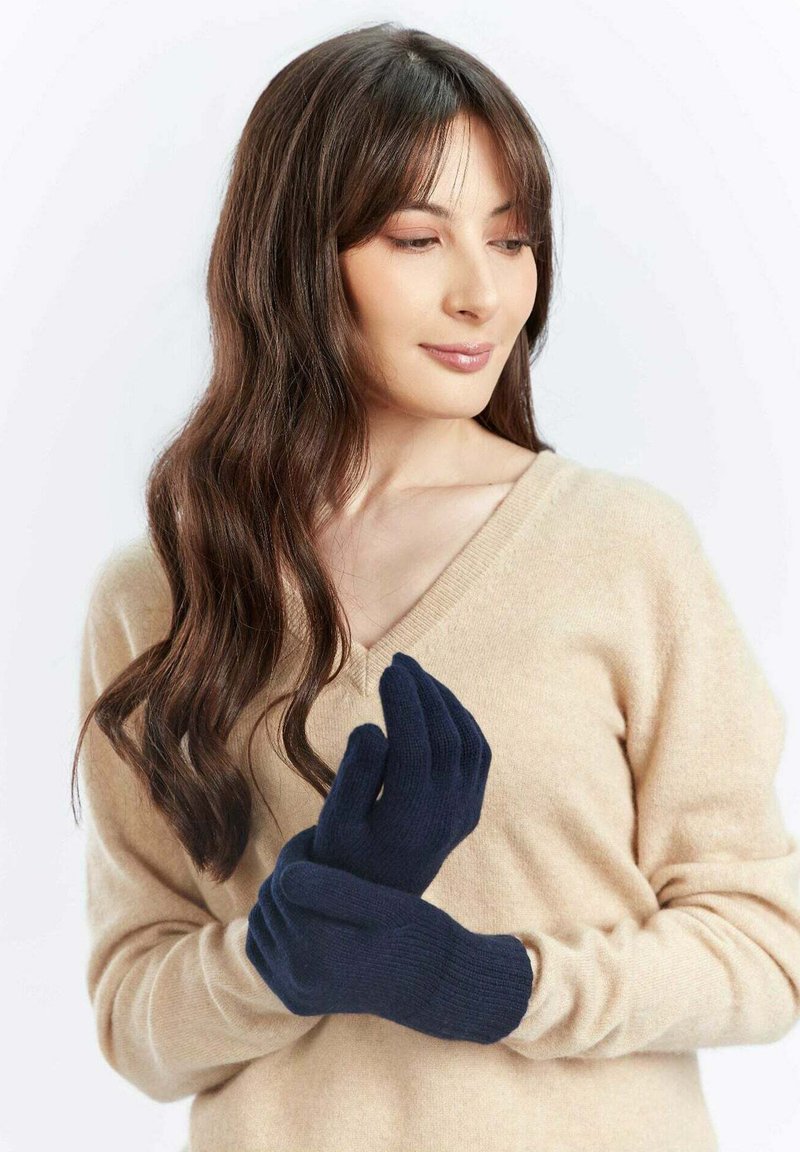 Marinblå stickade handskar med ribbad textur, med avsmalnande fingrar, som bärs av en person i en beige V-ringad tröja.