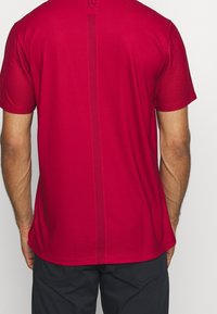 Röd atletisk t-shirt i något strukturerad tyg, med ett centralt vertikalt randmönster och korta ärmar.