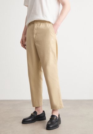 Osoba ubrana w beżowe spodnie o prostych nogawkach i czarne mokasyny, stojąca na jasnoszarej podłodze przy jednolitej białej ścianie.