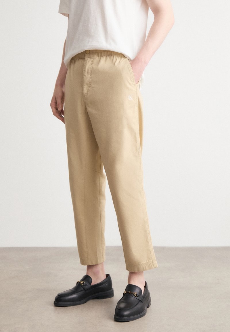 Personne portant un pantalon beige à jambes droites et des mocassins noirs, debout sur un sol gris clair devant un mur blanc uni.