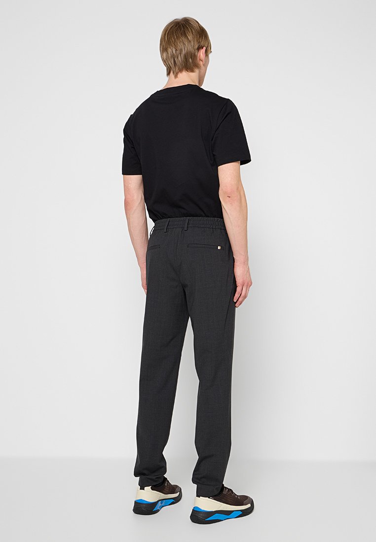 Mann in schwarzem T-Shirt, dunkelgrauen Hose mit elastischem Bund und schwarzen Turnschuhen mit weißen und blauen Sohle, von hinten stehend.