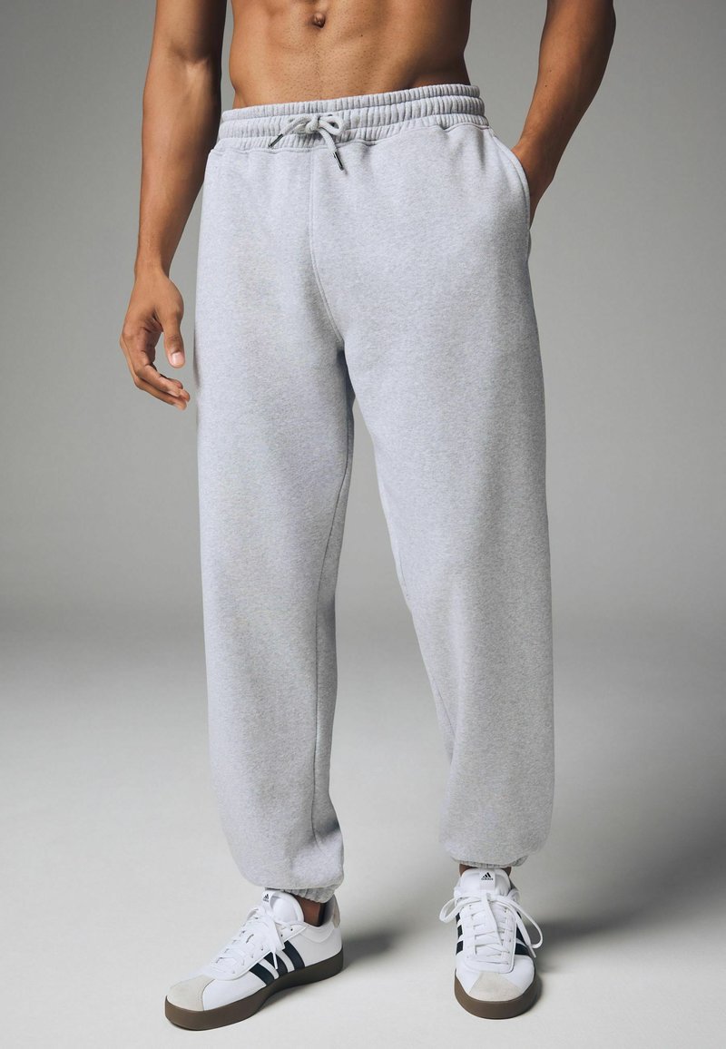 Graue Sweatpants aus weichem Material mit elastischem Bund und Kordelzug; an den Bündchen schmal zulaufend. Kombiniert mit weißen Sneakers mit schwarzen Streifen.