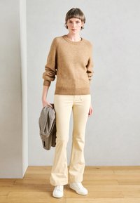 Hellbrauner Strickpullover mit langen Ärmeln, kombiniert mit cremefarbenen Schlaghosen und weißen Sneakers. Hält eine schwarz-weiße kariertes Jacke.