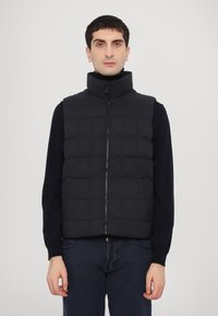 Black padded vest with a high collar, subtle quilted design, front zipper, worn over a dark long-sleeve top, paired with dark pants.