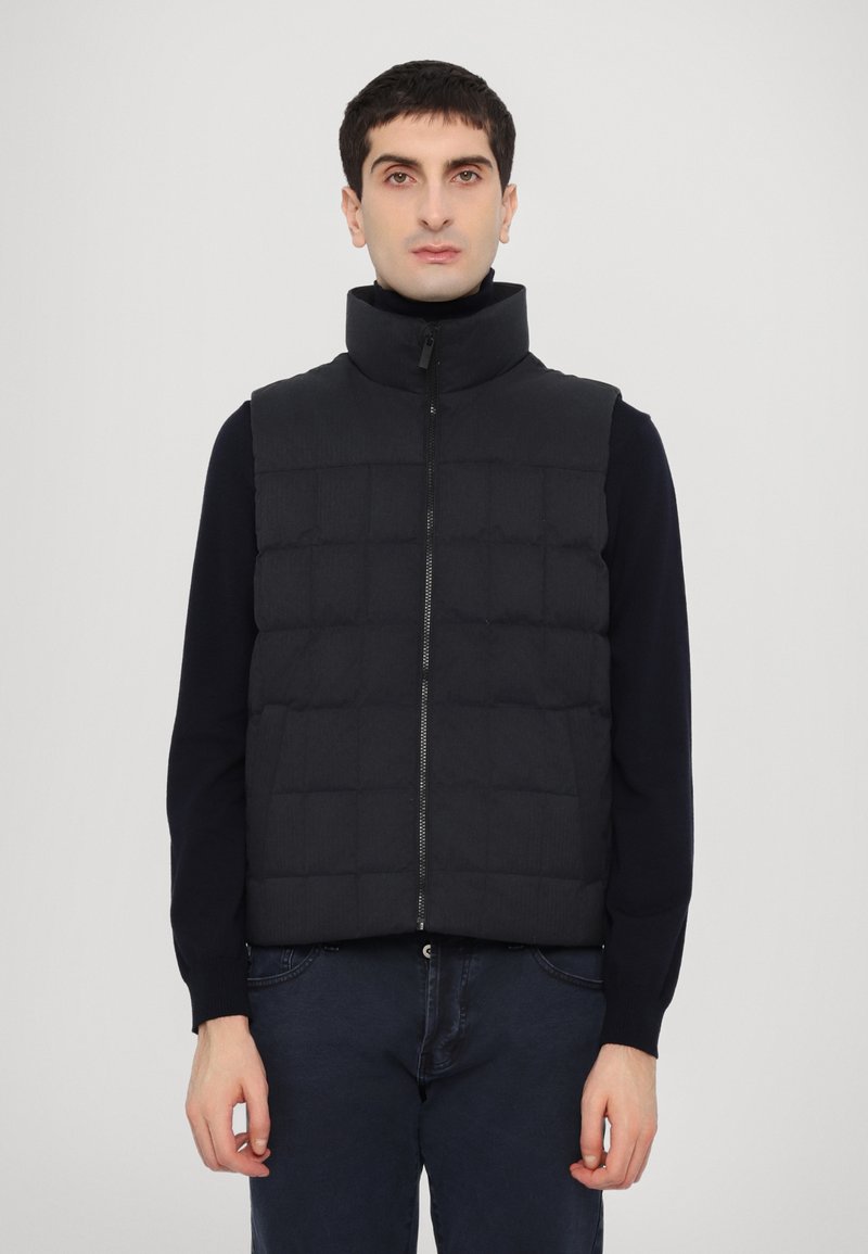 Black padded vest with a high collar, subtle quilted design, front zipper, worn over a dark long-sleeve top, paired with dark pants.