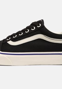 Czarne płócienne trampki z białym skórzanym paskiem po boku, czarnymi sznurówkami i fioletowym akcentem na podeszwie. Teksturowana gumowa podeszwa zapewniająca przyczepność.