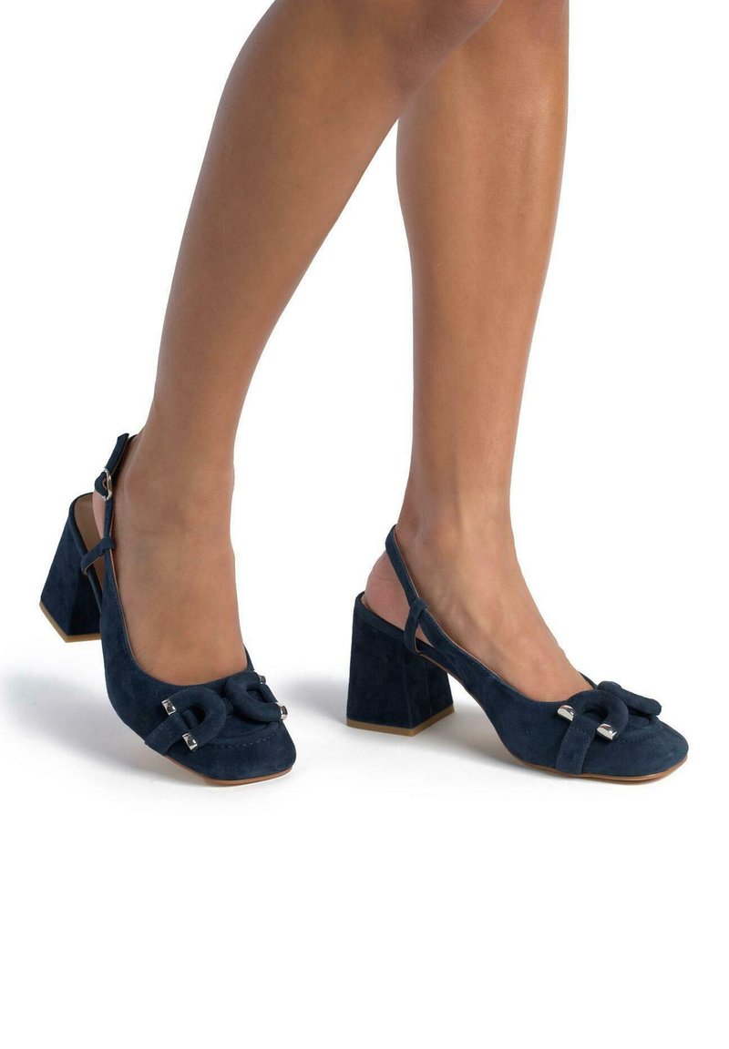 Granatowe zamszowe czółenka na odkrytej pięcie z kwadratowym noskiem, teksturowaną kokardą z przodu oraz grubym, wysokim obcasem. Zawiera akcenty w srebrnym odcieniu.