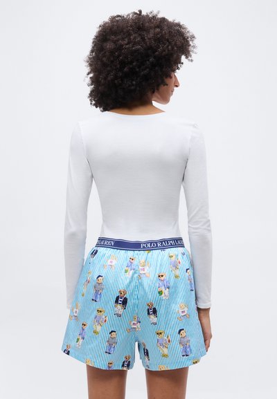 Γυναίκα με σγουρά μαλλιά που φοράει λευκό μακρυμάνικο τοπ και μπλε ριγέ σορτς με σχέδια αρκουδάκια και ζώνη μέσης Polo Ralph Lauren.