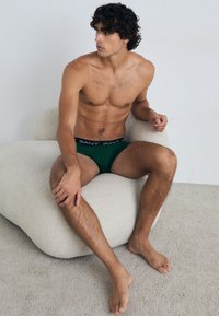 Bañadores verdes para hombre con una cinturilla negra de GANT, usados por un modelo masculino sentado sobre una superficie texturizada suave y de color claro.