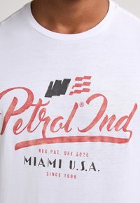 Bílá bavlněná tričko s červeným a černým vintage stylem textu "Petrol Ind" a "Miami U.S.A." s měkkou texturou a kulatým výstřihem.