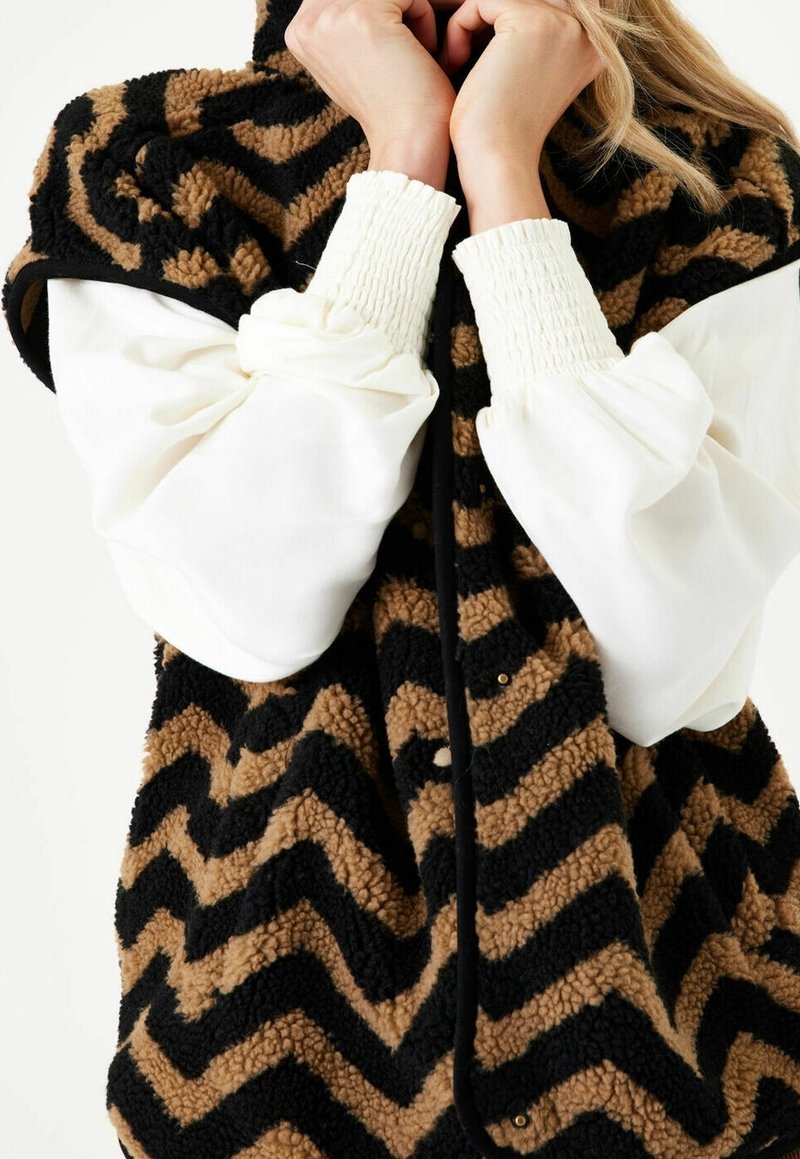 Faux fur-väst i brunt och svart zigzag-mönster, med en krage och tryckknappsstängning. Långa vita ärmar med elastiska manschetter synliga.