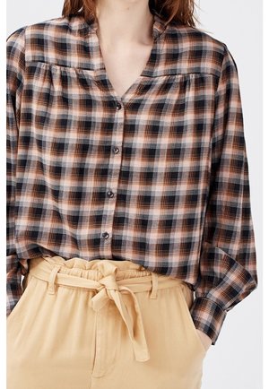 Vrouw draagt een zwart, bruin en wit geruit overhemd met knopen, ingestoken in beige paperbag taillebroeken met een gestrikte riem.