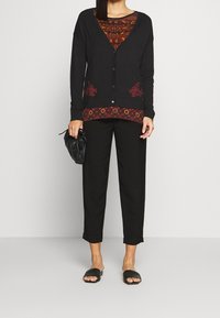Schwarze Strickjacke mit V-Ausschnitt, Knöpfen und roter Blumenstickerei am Saum. Über einer gemusterten Bluse getragen und kombiniert mit schwarzen Hosen und Sandalen.