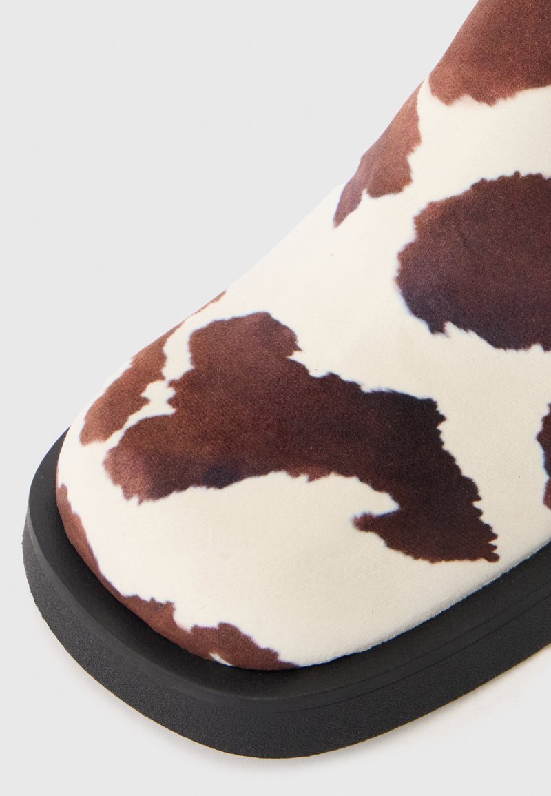Brown and white cow print ankle boot with a soft texture, rounded toe, and a black rubber sole. Close-up view highlights the design detail.