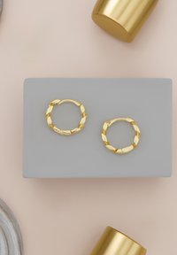 Boucles d'oreilles créoles en or avec un design torsadé, présentées sur une plateforme grise. La texture lisse et la finition brillante mettent en valeur leur apparence.