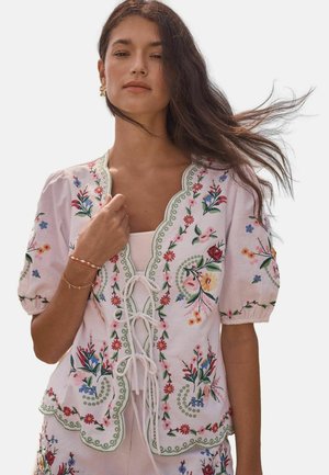 Kobieta w białej bluzce z falistymi krawędziami i kolorowym kwiatowym haftem, związana na przodzie koronką, jej włosy delikatnie powiewają do tyłu.