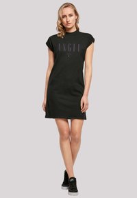 Schwarzes Baumwollkleid mit Rundhalsausschnitt, kurzen Ärmeln und einem lila "ANGEL"-Grafikdesign auf der Vorderseite. Getragen mit schwarzen High-Top-Sneakern.