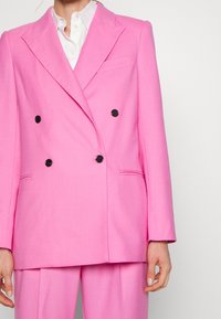 Blazer doppiopetto in tessuto rosa brillante, con bottoni neri, reverse strutturati e tasche laterali. Presenta una texture liscia e una vestibilità sartoriale.