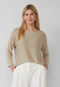Beige Strickpullover mit langen Ärmeln und lockerer Passform. Der Stoff hat einen subtilen Schimmer. Kombiniert mit weißen Hosen, die einen leichten Kontrast schaffen.