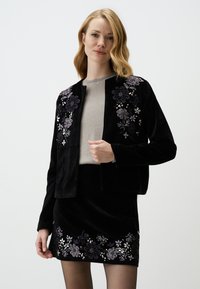 Giacca di velluto nero con ricami floreali in viola e argento, indossata sopra un top grigio chiaro e abbinata a una gonna coordinata.