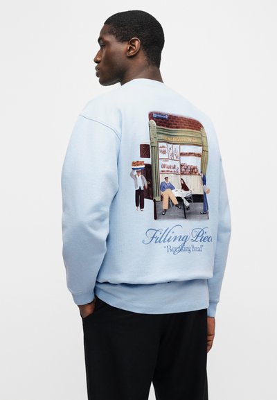 Άντρας που φοράει φωτεινό μπλε φούτερ με εικονογράφηση πρόσοψης φούρνου και ανθρώπων, με το κείμενο «Filling Pieces 'Breaking Bread'» στην πλάτη.