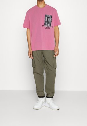Pink cotton t-shirt with a black and white graphic print, paired with olive green cargo pants and white sneakers. Casual and relaxed fit.