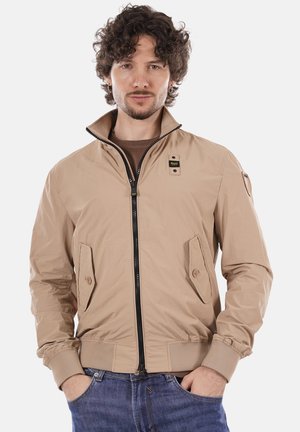 Uomo con capelli ricci che indossa una giacca beige con zip e jeans blu, mani nelle tasche, sfondo neutro.