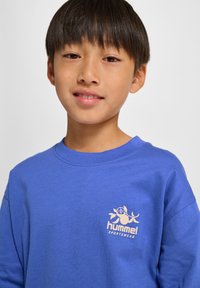 Blå bomullströja med rund halsringning, långa ärmar och en liten beige logga på vänstra bröstet. Mjuk textur.