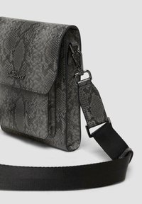 Szara torebka typu crossbody w wężowy wzór z zapięciem na klapkę, z teksturowaną powierzchnią, regulowanym czarnym paskiem oraz detalami logo z przodu.