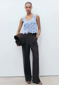Top smanicato blu all'uncinetto con motivi floreali, abbinato a pantaloni marroni a vita alta. La modella tiene una borsa a maglia marrone scuro. Texture liscia.