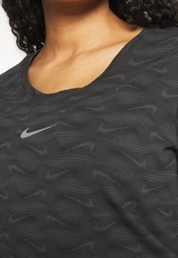 Svart tränings T-shirt med ett vågmönster och en ljusgrå Nike swoosh-logotyp. Rund halsringning och textilier med struktur.