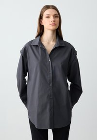 Chemise boutonnée gris foncé avec un col structuré, des manches longues et des détails de poignets retroussés. Tissu lisse avec une coupe décontractée.